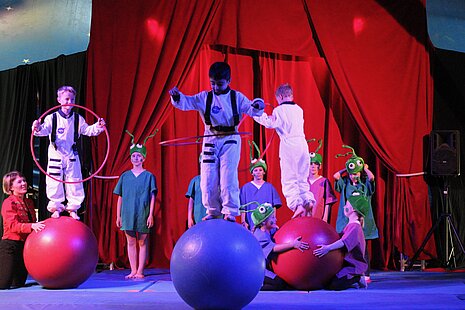 Schülerinnen und Schüler balancieren in einer Zirkusmanege auf großen Bällen